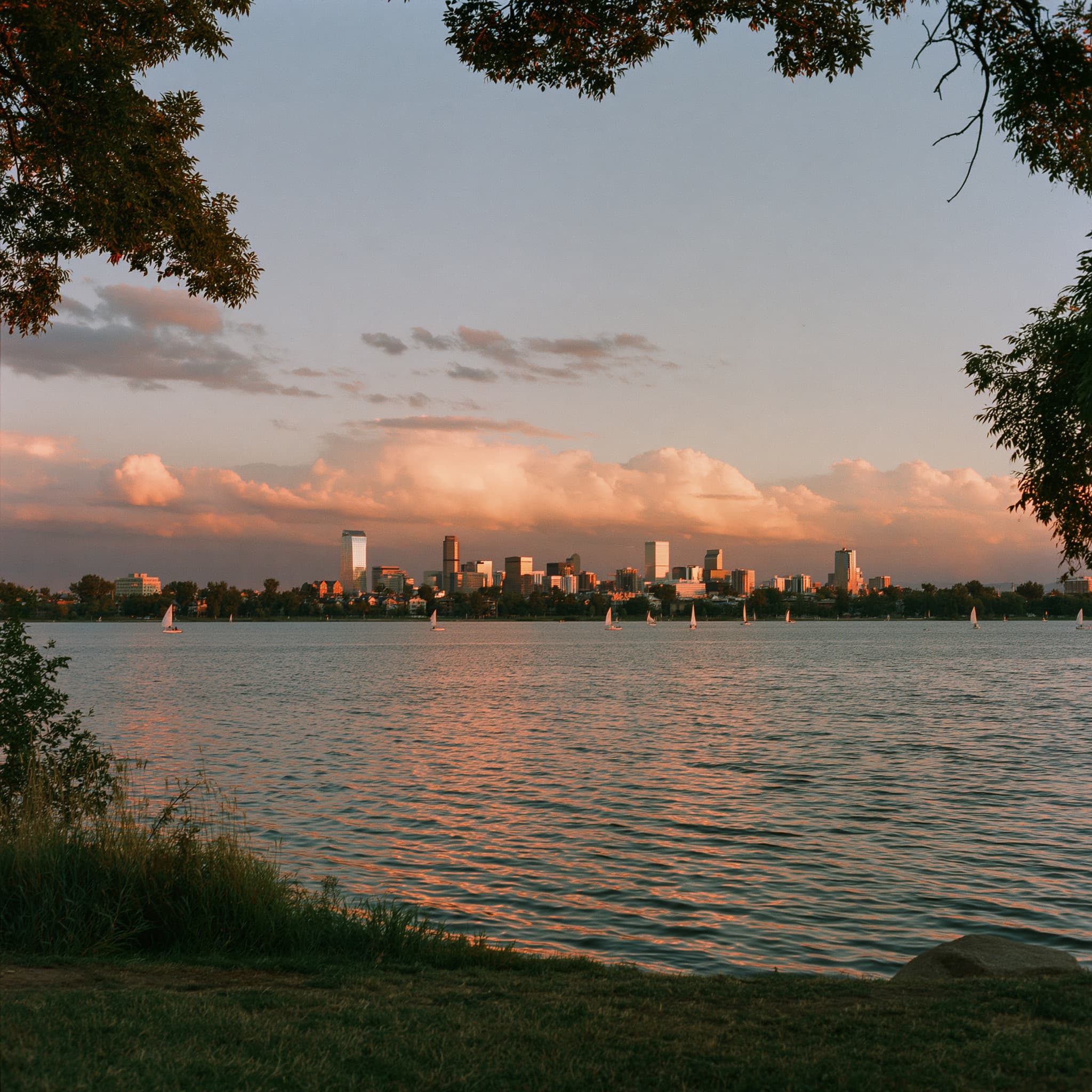 A wide lake at sunset with the Denver skyline low on the far shore and sailboats on the water